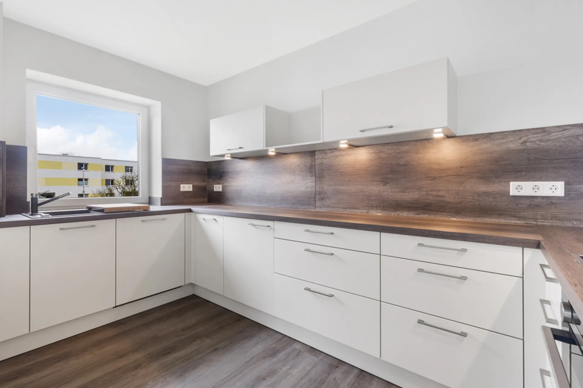 Moderne Einbauküche mit weißen Schränken, Holzarbeitsplatten und großem Fenster.