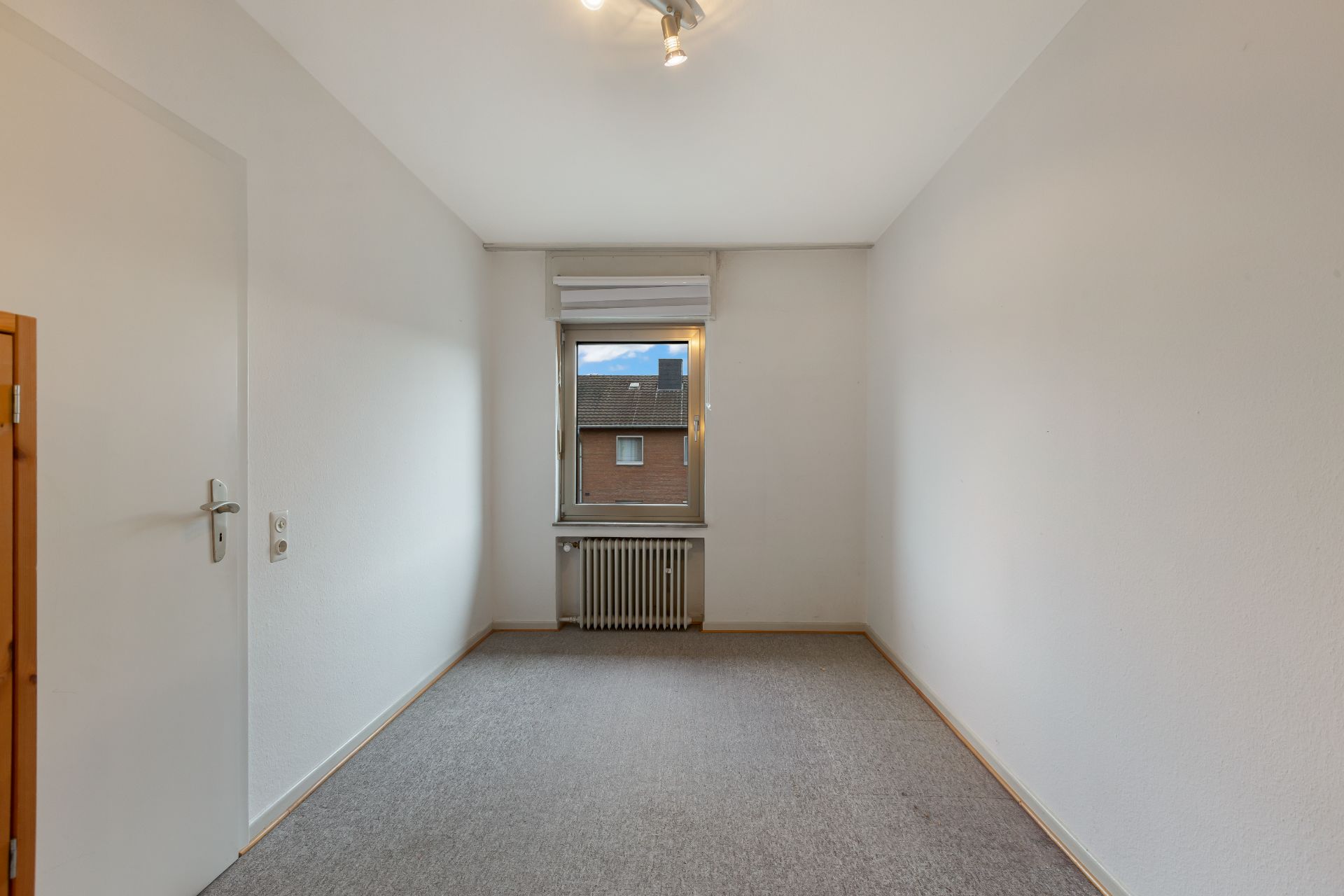 Leerer Raum mit Teppichboden, weiß gestrichenen Wänden und einem Fenster mit Blick nach draußen.