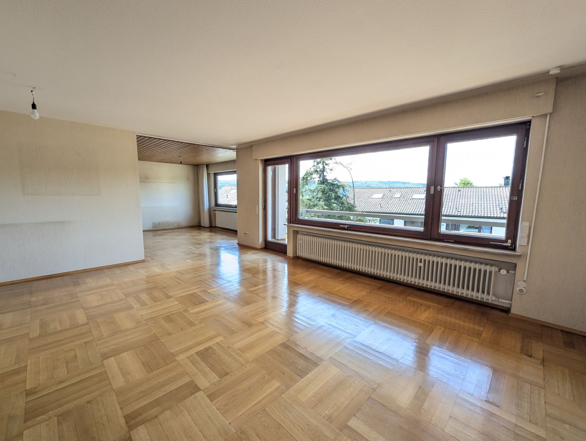 Leeres Wohnzimmer mit Parkettboden und großen Fenstern, Blick auf Dächer und Bäume.