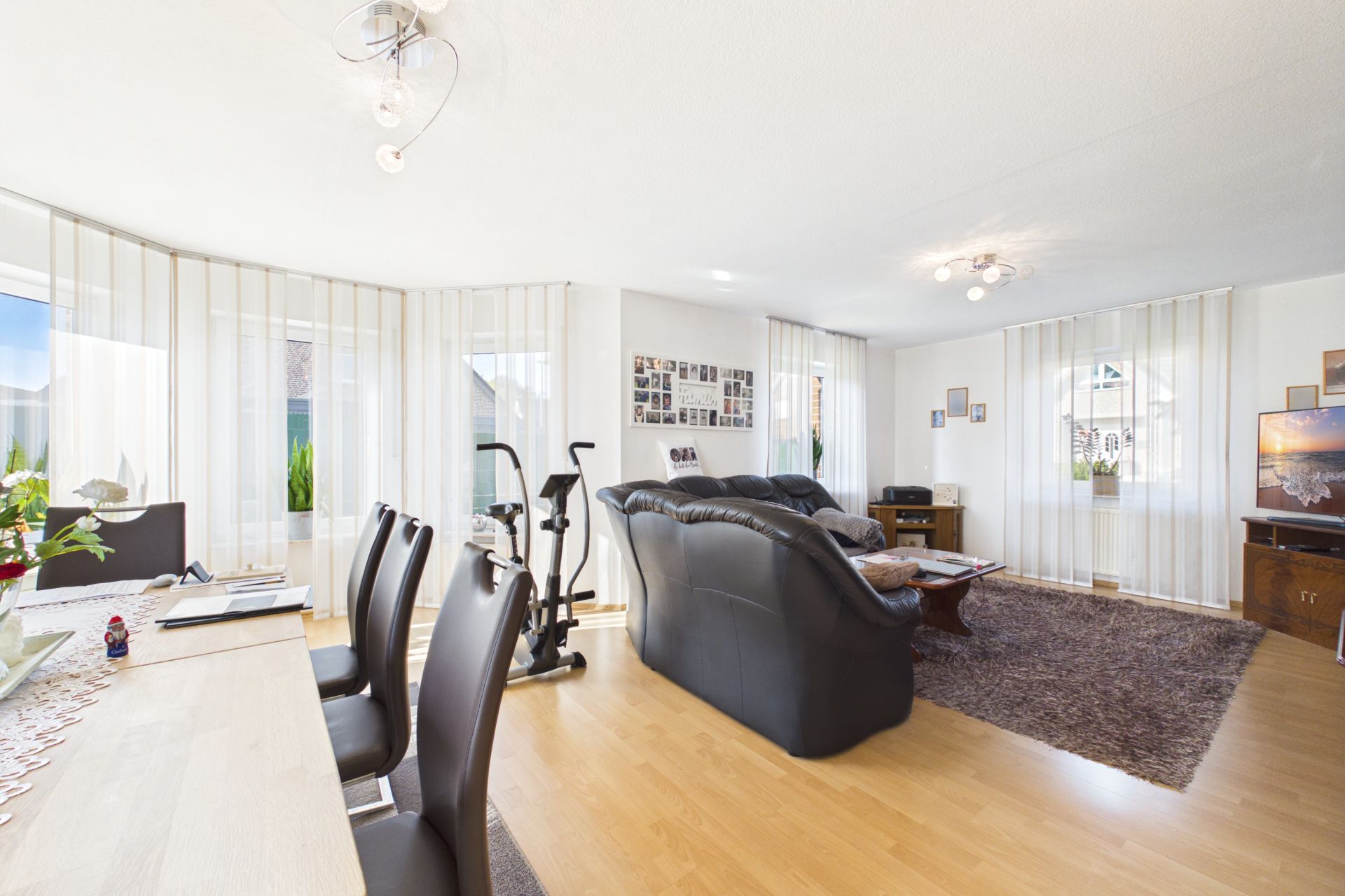 Helles Wohnzimmer mit schwarzem Sofa, Esstisch, Teppich und Heimtrainer auf Holzfußboden.