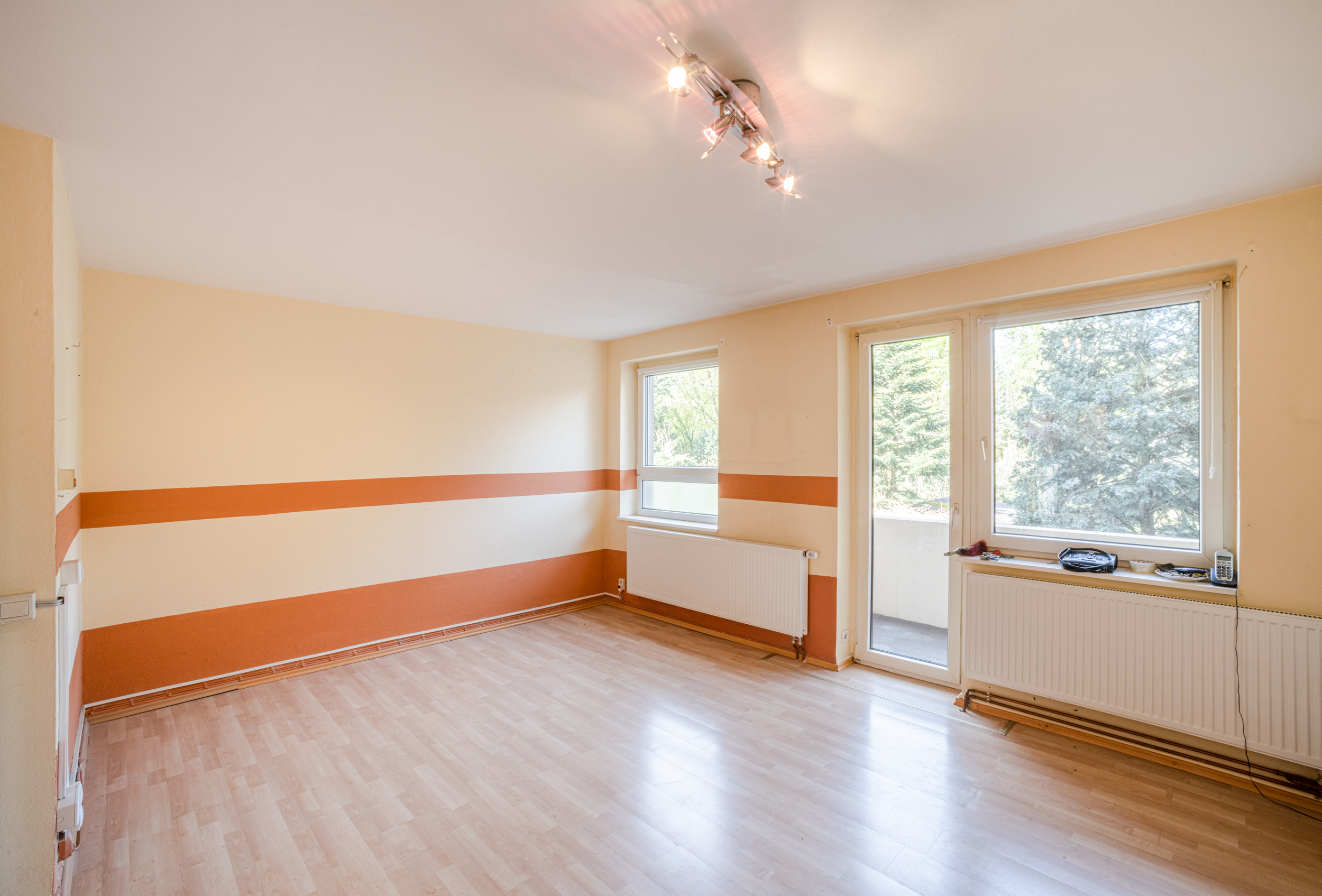 Helles, leeres Zimmer mit Holzfußboden, großen Fenstern und orangefarbenen Streifen an den Wänden.