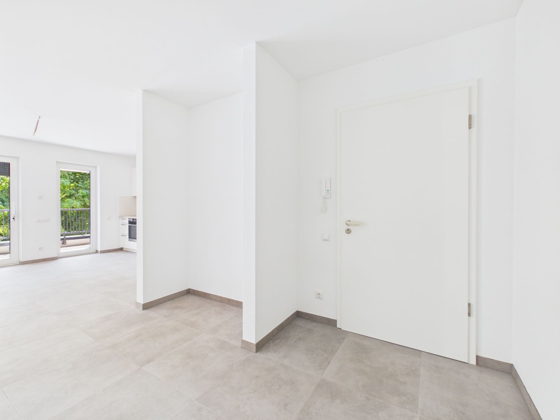 Heller Eingangsbereich mit weißer Tür, Blick ins Wohnzimmer mit Fenstern und Balkon.