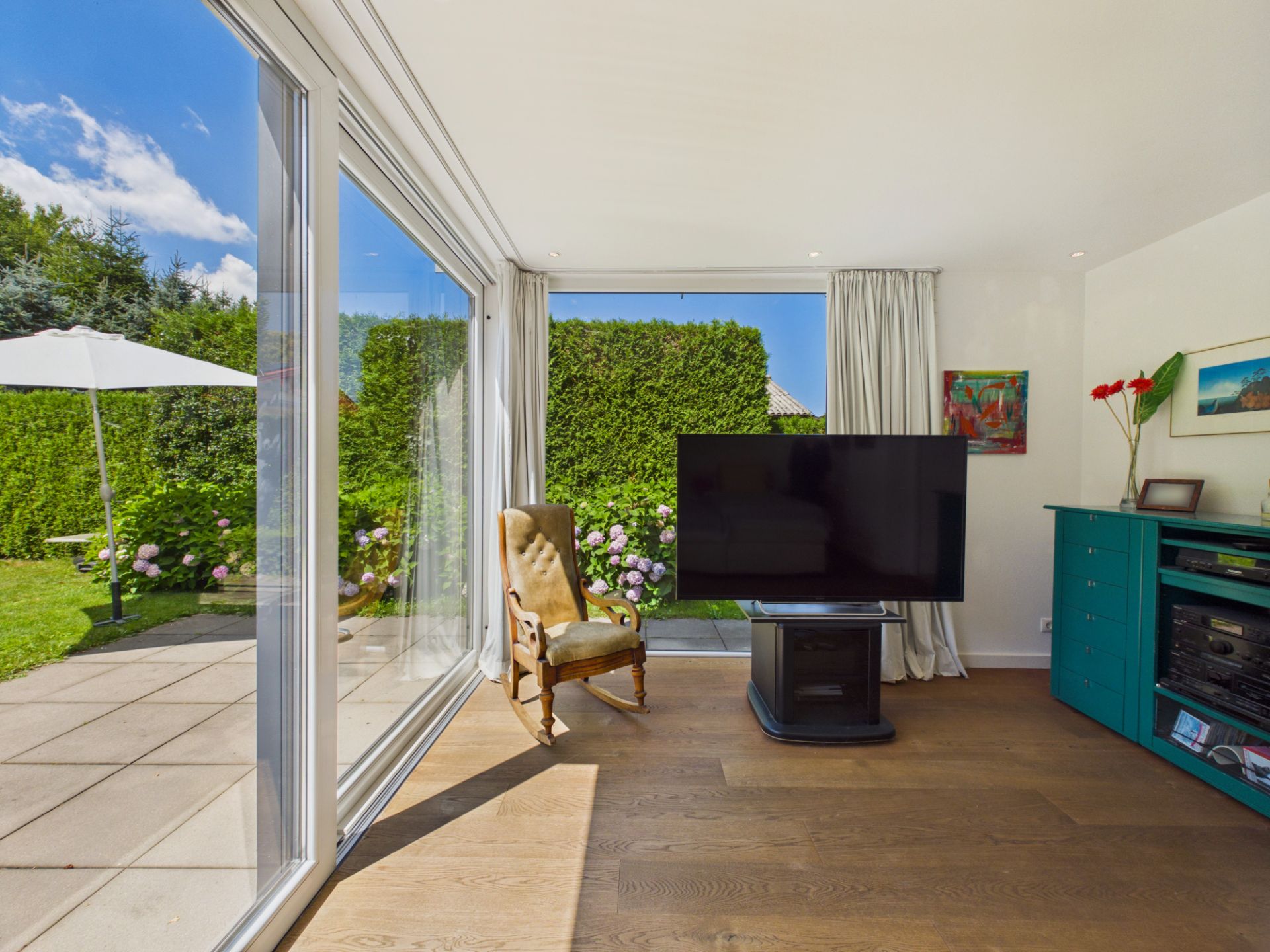 Wohnzimmer mit großem Fenster, Sessel, Fernseher und Blick in den Garten mit Sonnenschirm.