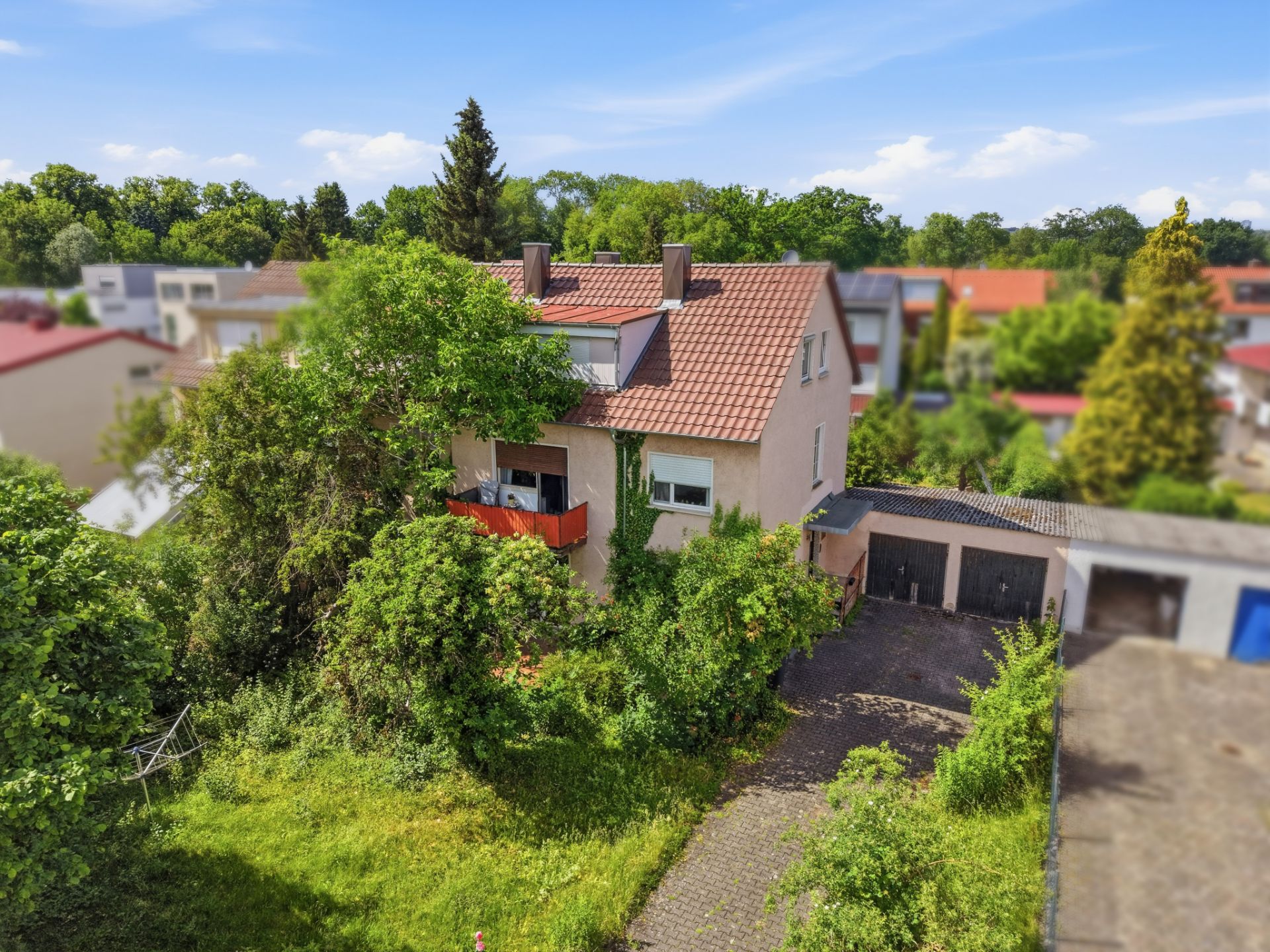 Zweistöckiges Wohnhaus mit rotem Balkon, Garten und Garagen, umgeben von Bäumen.