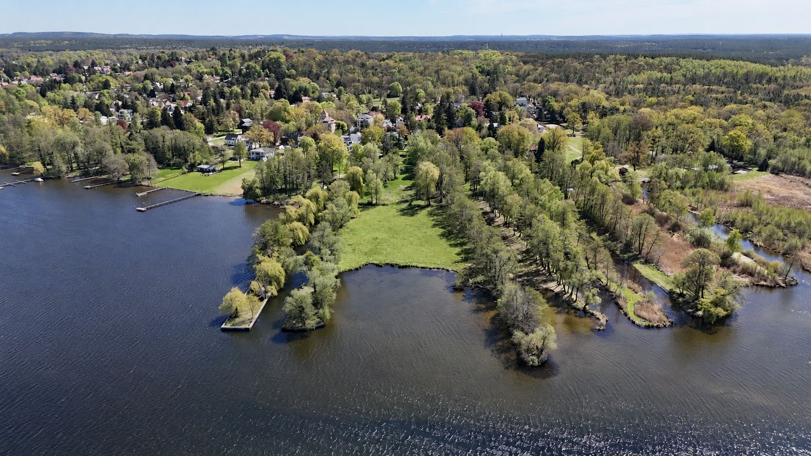 Luftaufnahme einer bewaldeten Halbinsel mit Wiesen am See und angrenzender Wohnsiedlung.