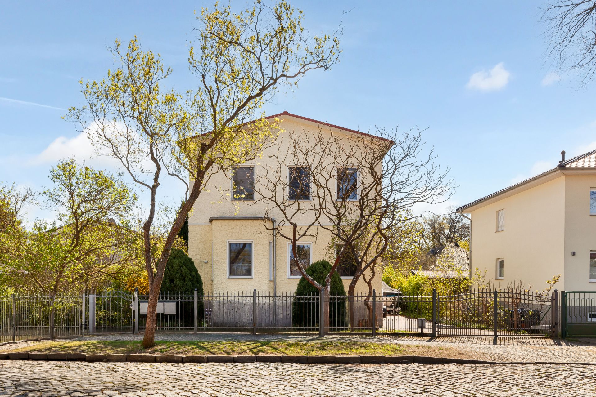 Zweistöckiges Wohnhaus mit gelber Fassade, Bäumen im Vorgarten und Zaun an einer gepflasterten Straße.