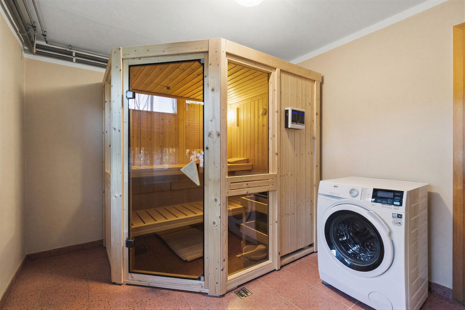 Holzsauna mit Glastür neben einer weißen Waschmaschine in einem hellen Raum.