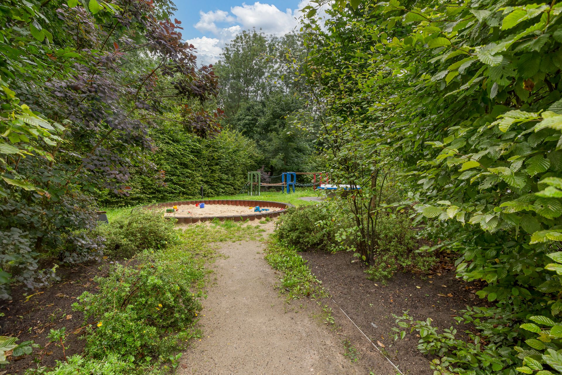 Gartenbereich mit Sandkasten, umgeben von Sträuchern und Bäumen, Spielgeräte im Hintergrund.