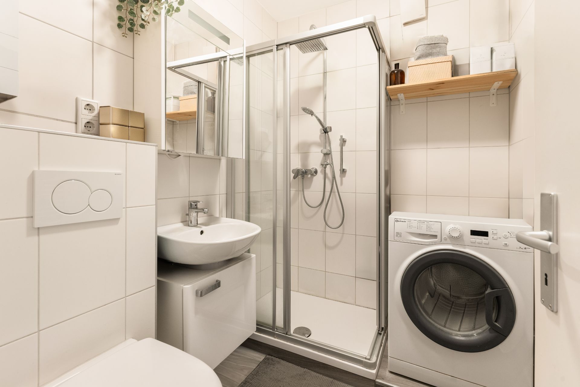 Modernes Bad mit Dusche, Waschbecken, Waschmaschine und Regal auf weiß gefliesten Wänden.