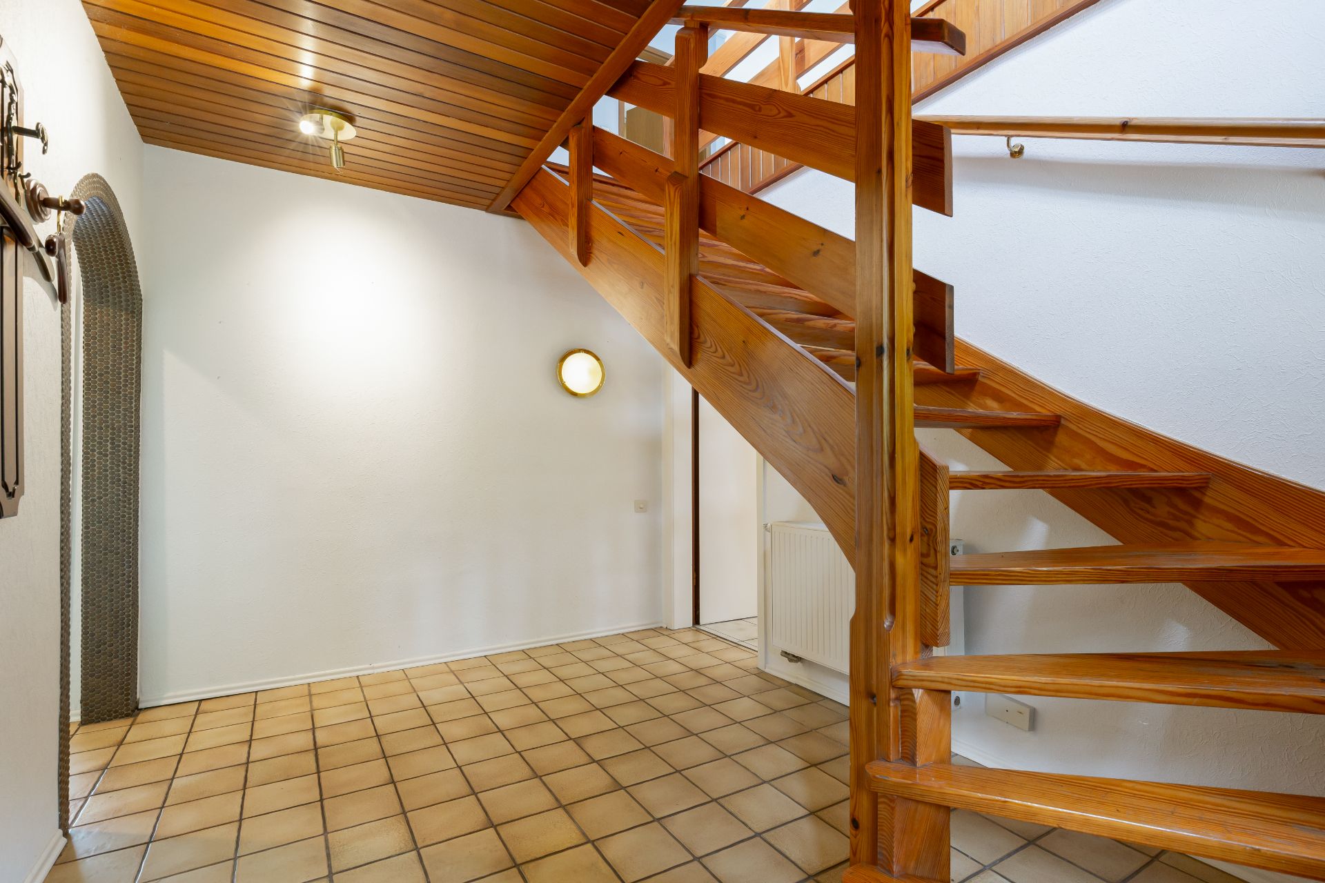 Flur mit gefliestem Boden und hölzerner Treppe, weiße Wände und Holzdecke.