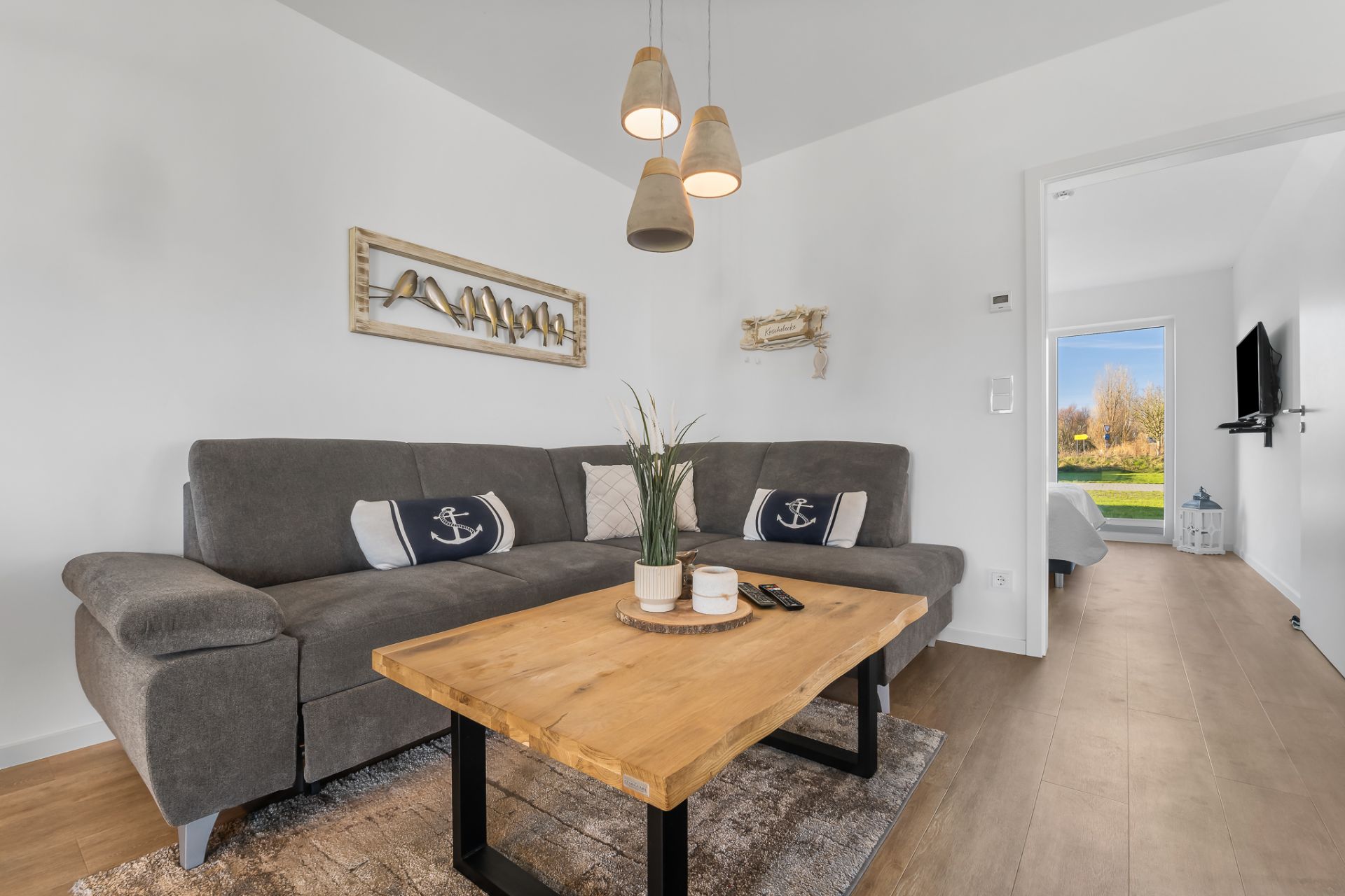 Wohnzimmer mit grauem Sofa, Holztisch, Deko-Kissen und Wandbildern, Tür zum Schlafzimmer.