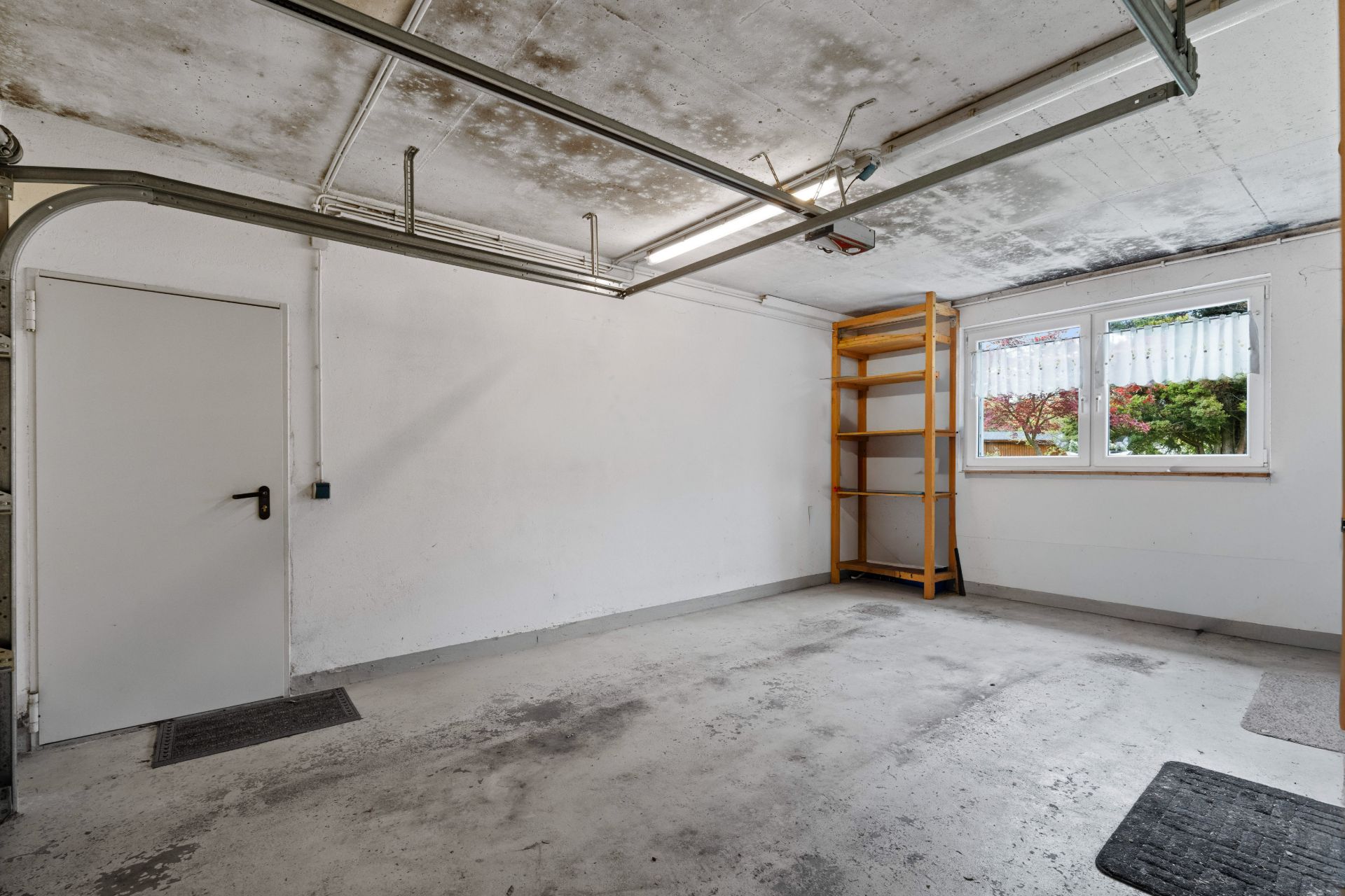 Leere Garage mit Regal, Tür links und zwei Fenstern rechts.