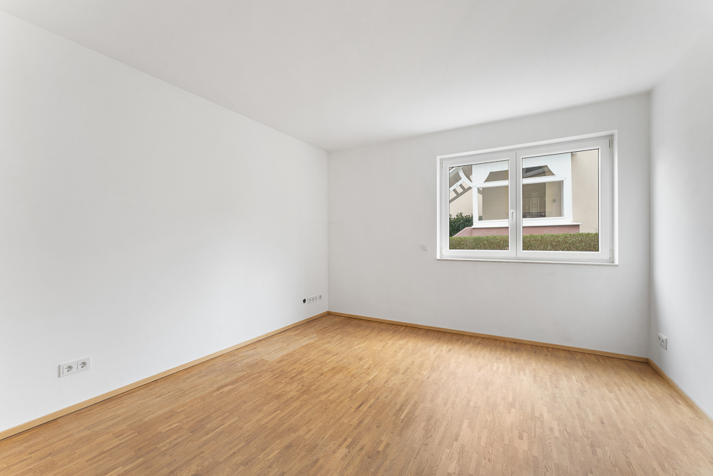 Leerer Raum mit hellem Holzboden und großem Fenster, weiße Wände.