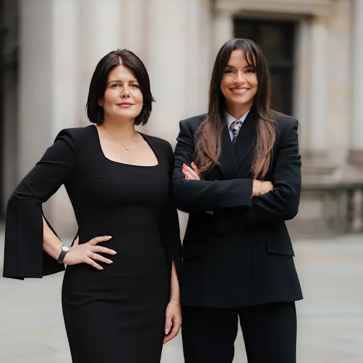 Nicole Herbst und Shpresa Achenbach stehen in eleganter Businesskleidung vor einem Sandsteingebäude und lächeln
