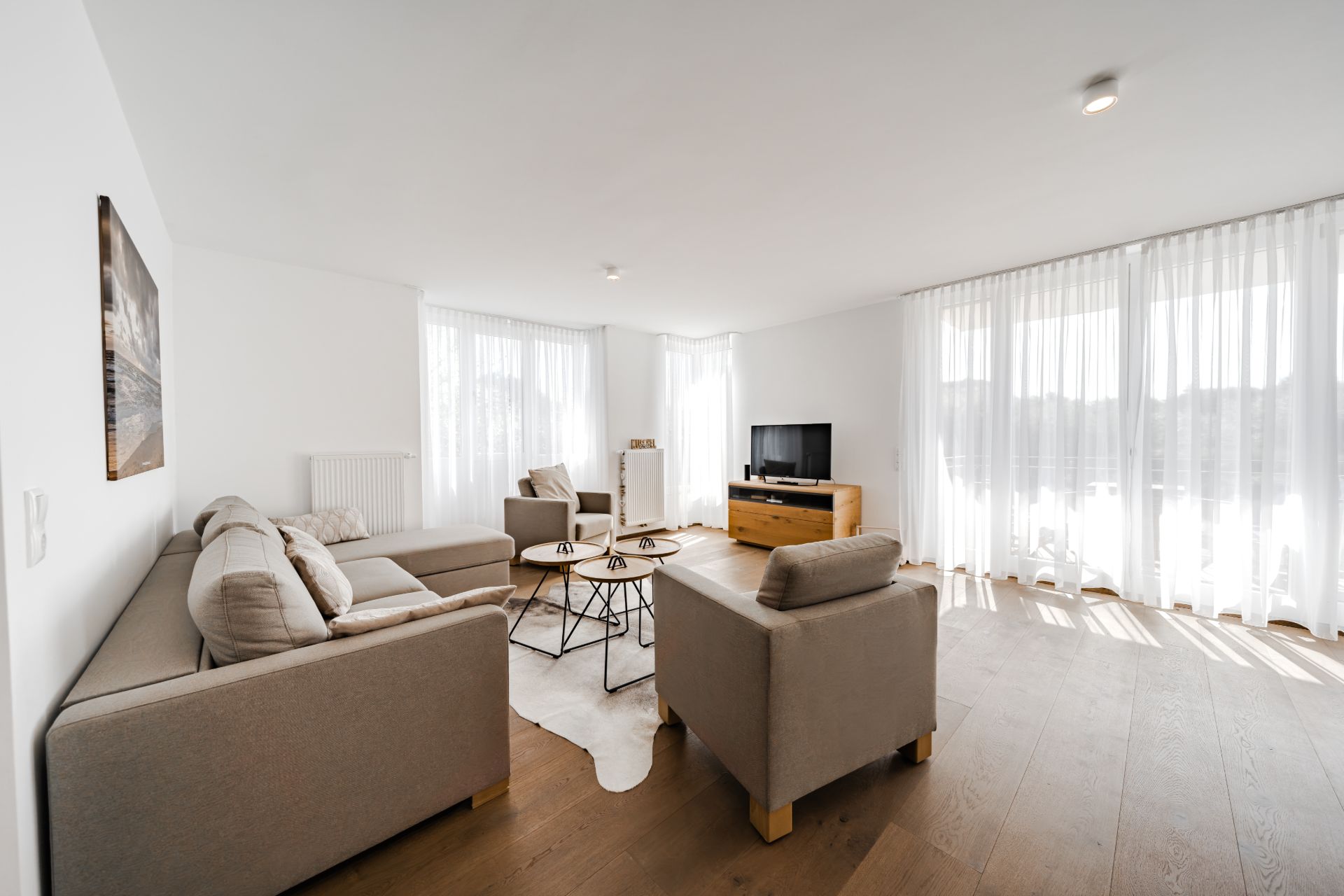 Helles Wohnzimmer mit Sofa, Sesseln, Couchtisch, Fernseher und großen Fenstern mit Vorhängen.