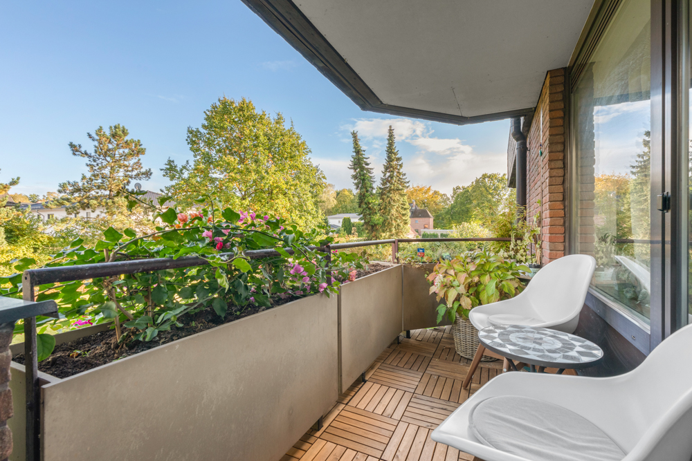 Balkon mit Holzboden, zwei weißen Stühlen, kleinem Tisch und Pflanzen, Blick auf Bäume.