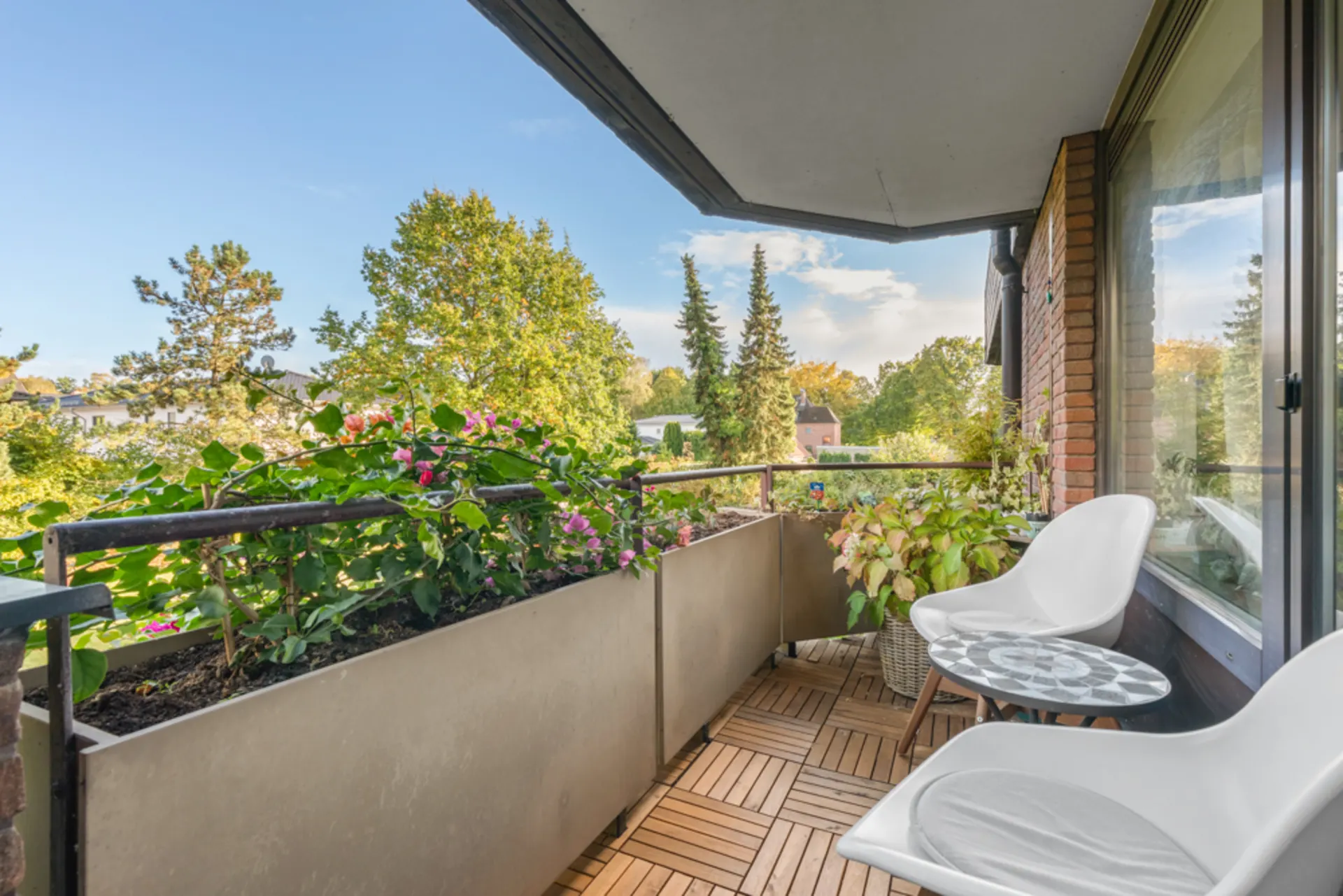 Balkon mit Holzboden, zwei weißen Stühlen, kleinem Tisch und Pflanzen, Blick auf Bäume.
