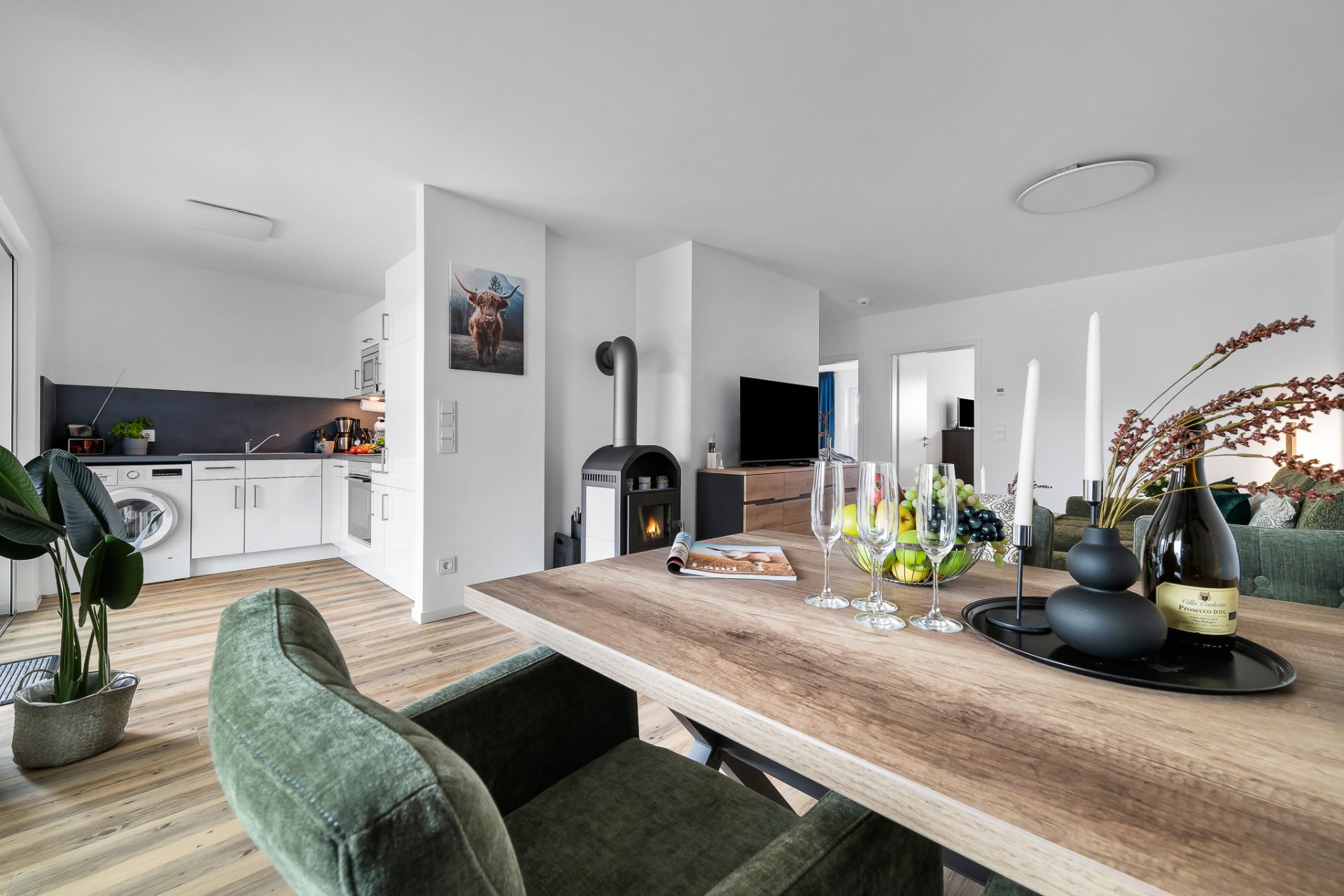 Helles Wohnzimmer mit Esstisch, Kaminofen, offener Küche und grüner Einrichtung.