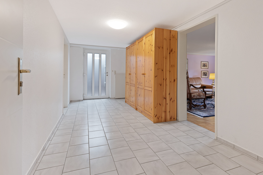 Heller Flur mit Fliesenboden, Holzschrankwand und Glastür, Zugang zu Wohnzimmer sichtbar.
