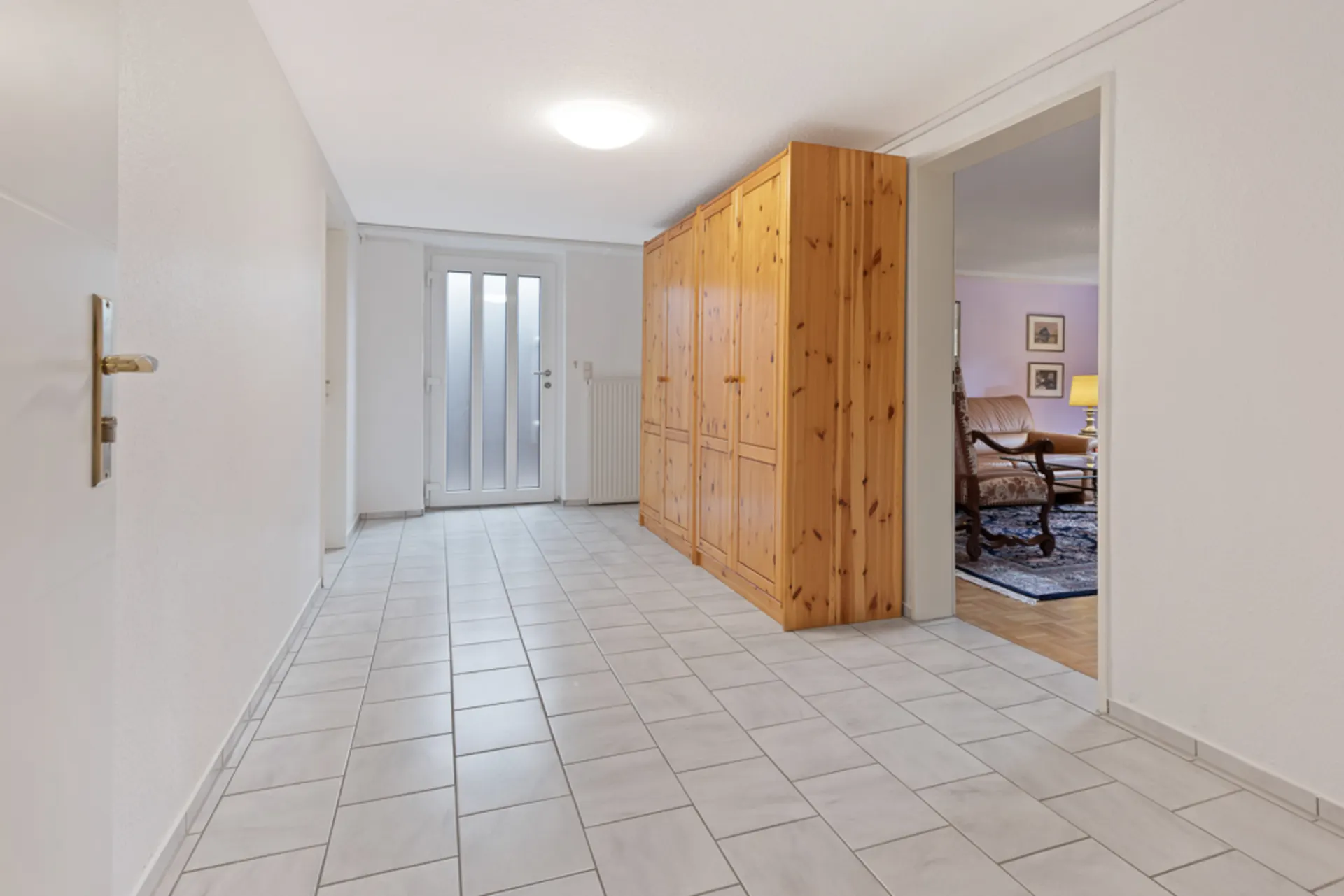 Heller Flur mit Fliesenboden, Holzschrankwand und Glastür, Zugang zu Wohnzimmer sichtbar.