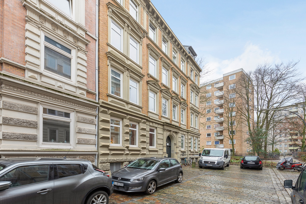 Mehrstöckiges Wohngebäude mit geparkten Autos auf einem gepflasterten Hof und weiteren Häusern im Hintergrund.