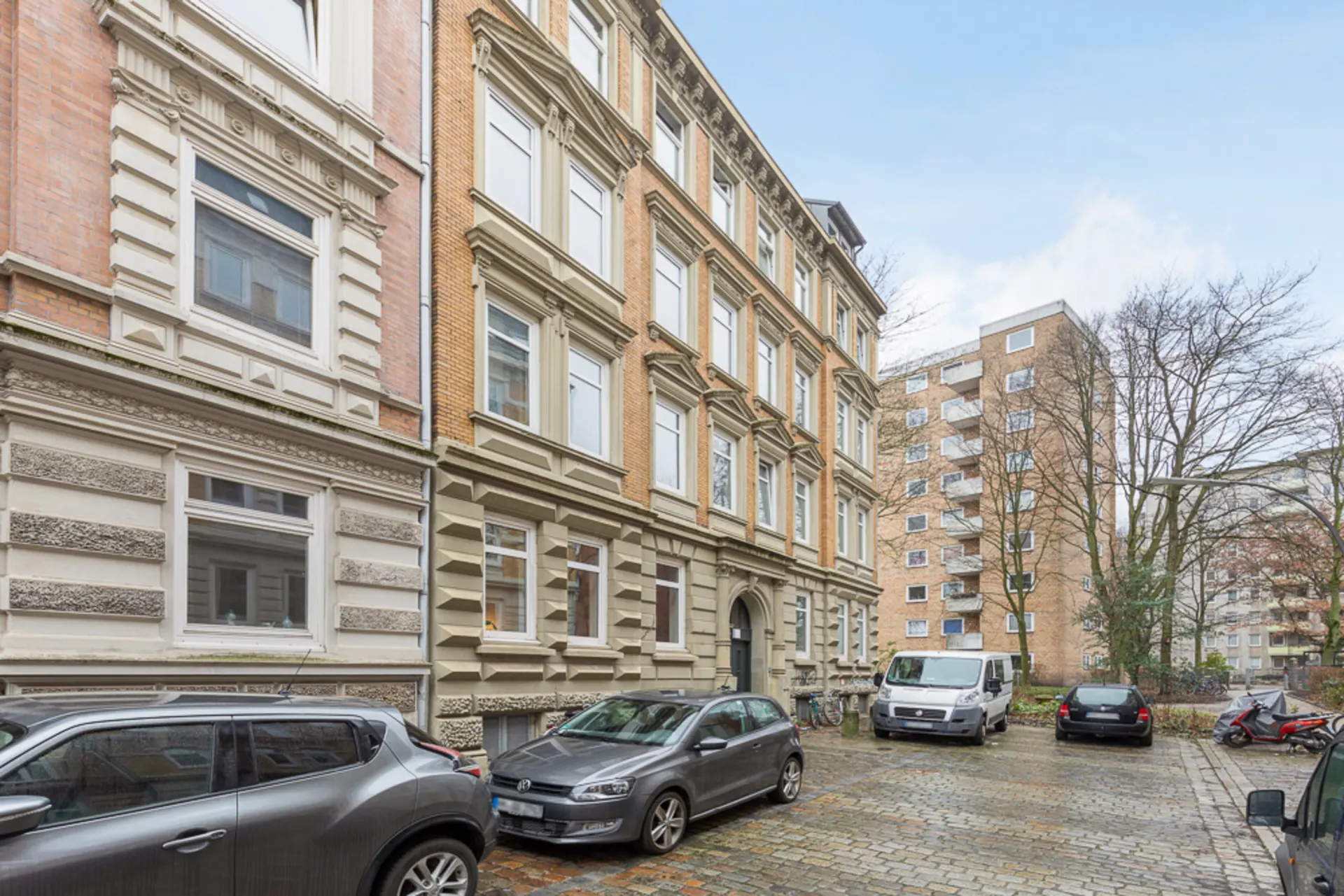 Mehrstöckiges Wohngebäude mit geparkten Autos auf einem gepflasterten Hof und weiteren Häusern im Hintergrund.