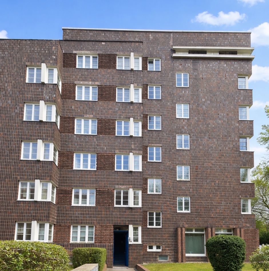 Mehrstöckiges Wohnhaus mit brauner Backsteinfassade und weißen Fenstern, Eingang unten mittig.
