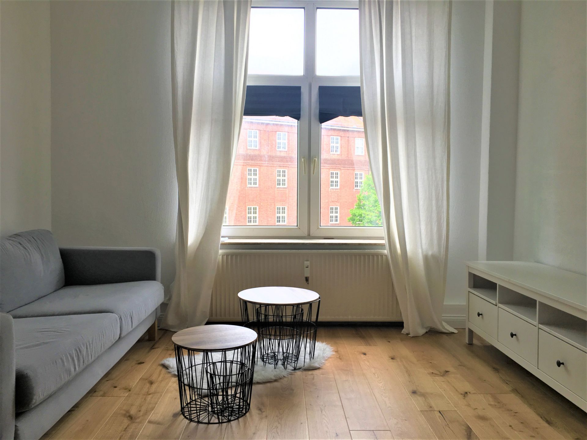 Helles Wohnzimmer mit grauem Sofa, zwei runden Tischen und großem Fenster mit weißen Vorhängen.