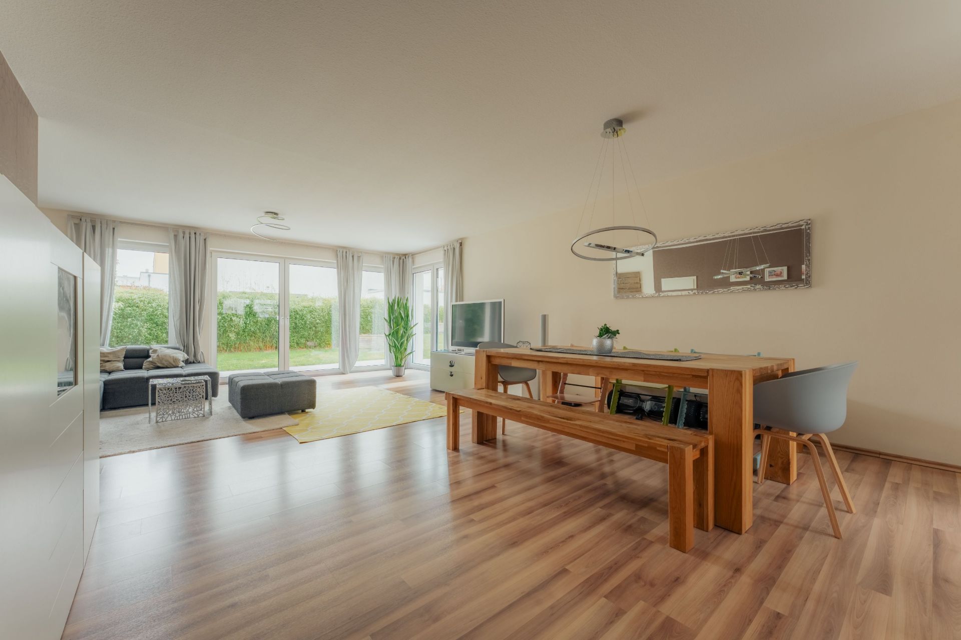 Helles Wohnzimmer mit Holzboden, Esstisch, Sofa, Fernseher und großen Fenstern zum Garten.