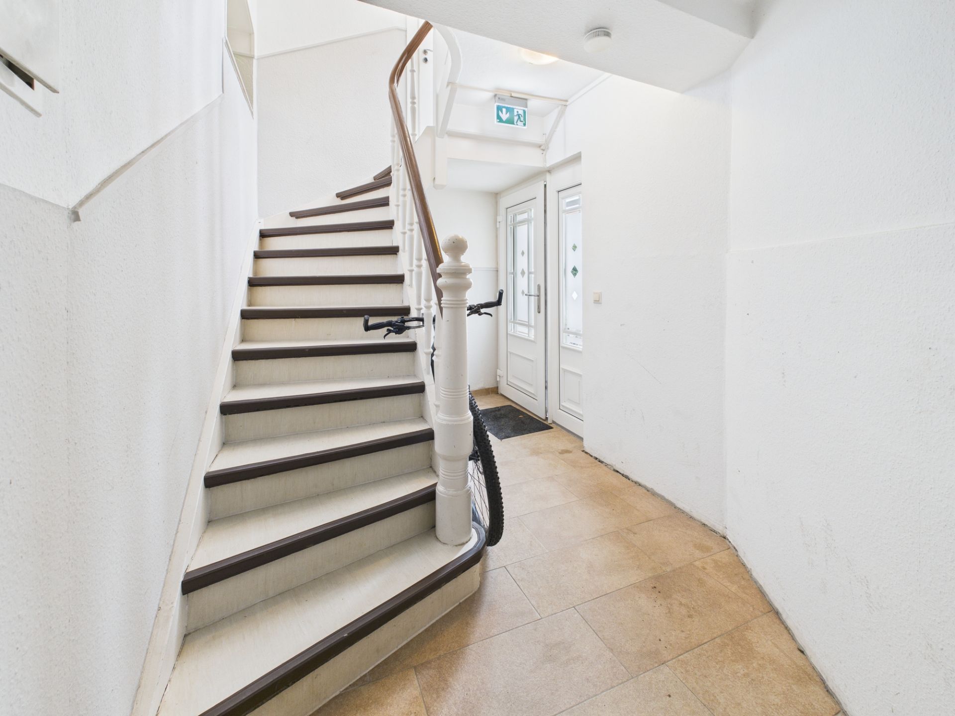 Helles Treppenhaus mit weißer Treppe, Fahrrad und zwei Glastüren im Eingangsbereich.