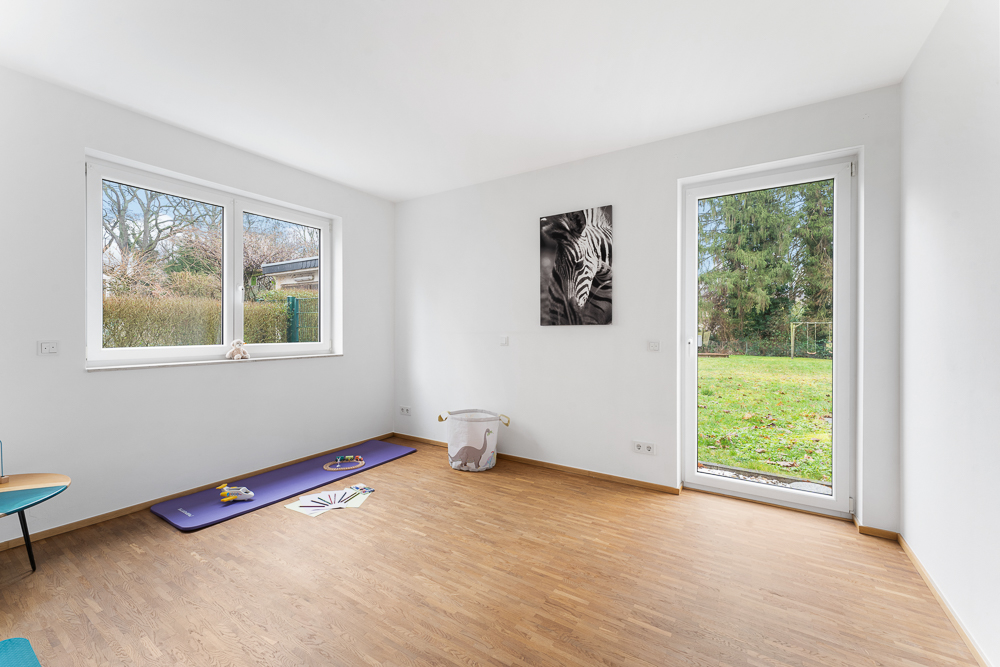 Heller Raum mit Holzfußboden, zwei Fenstern, Tür zum Garten und Spielzeug auf einer Matte.