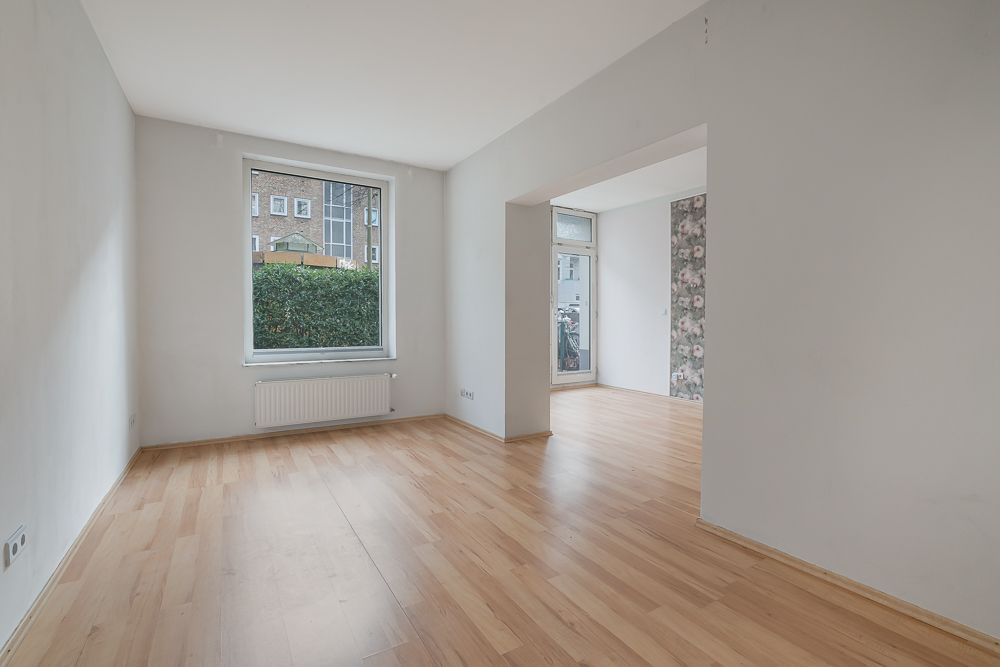 Helles, leeres Zimmer mit Holzfußboden, großem Fenster und Durchgang zu weiterem Raum.