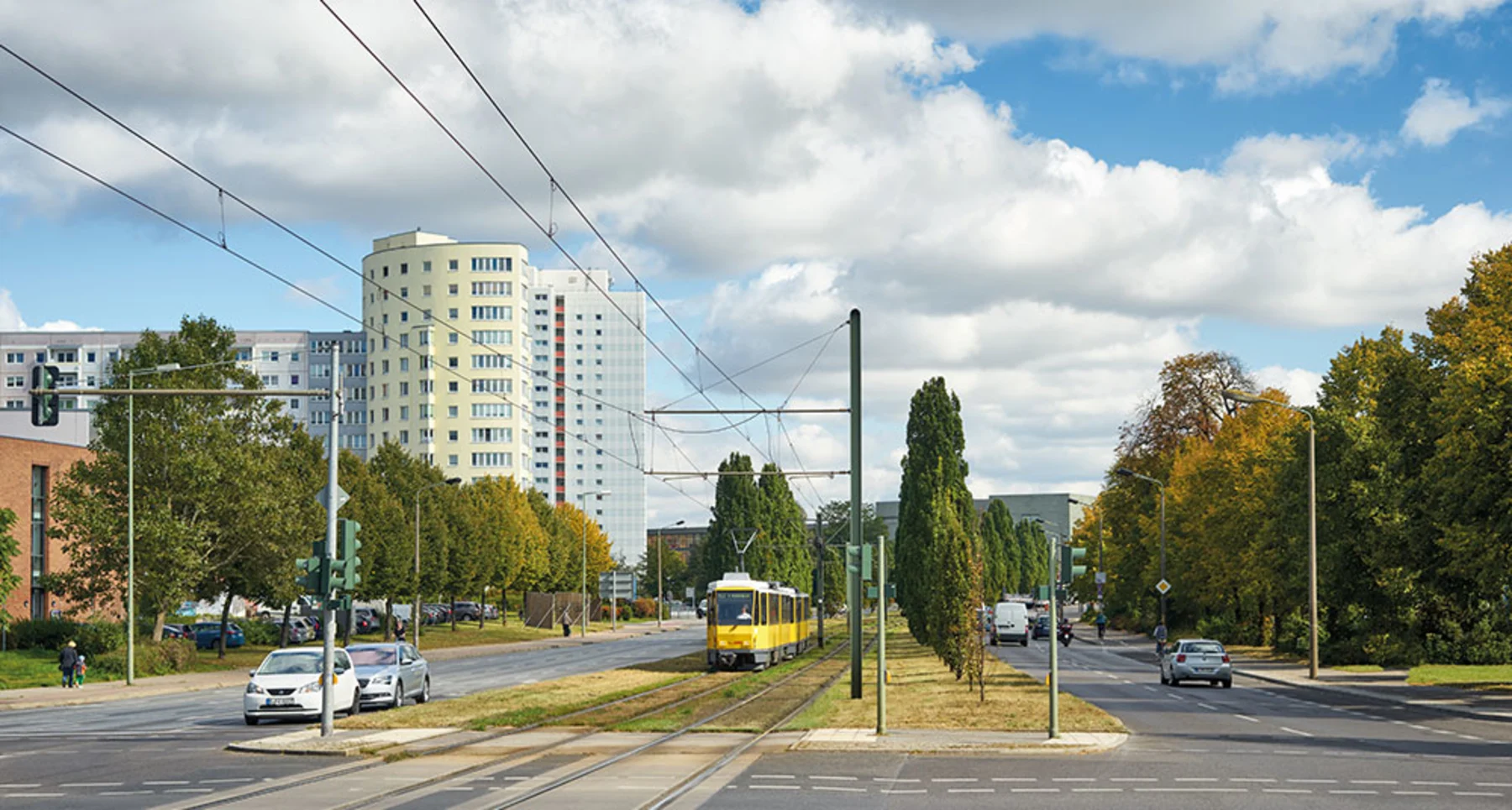 Wohngebäude im Hintergrund, Straßenbahn und Autos auf einer breiten Straße, Bäume an den Straßenrändern.
