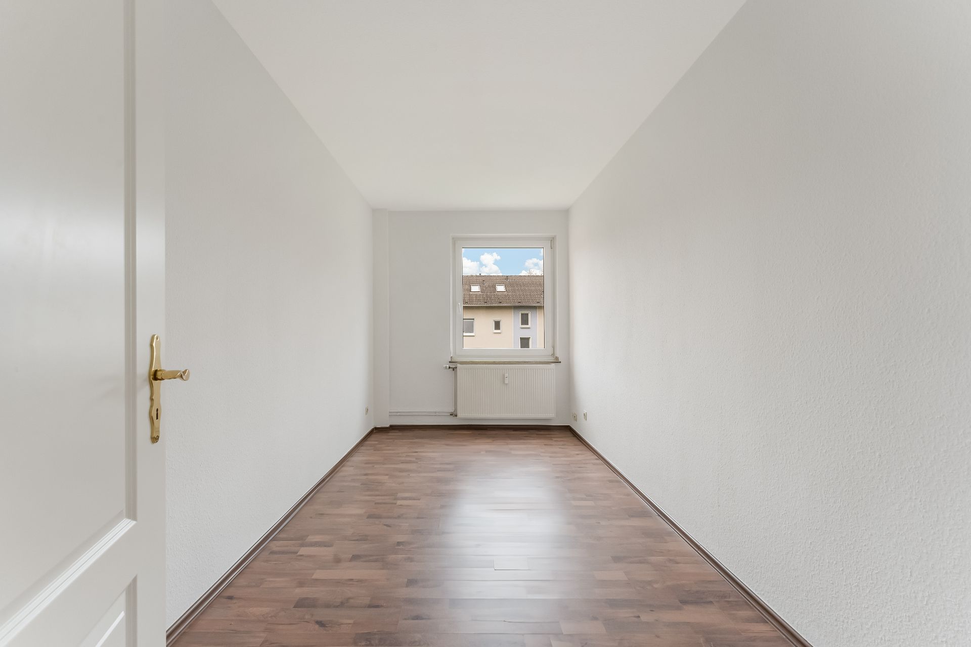 Leeres, helles Zimmer mit weißer Wand, braunem Boden und Fenster am Ende.