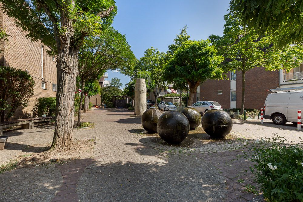 Gepflasterter Innenhof mit Bäumen, großen Steinkugeln und parkenden Autos.