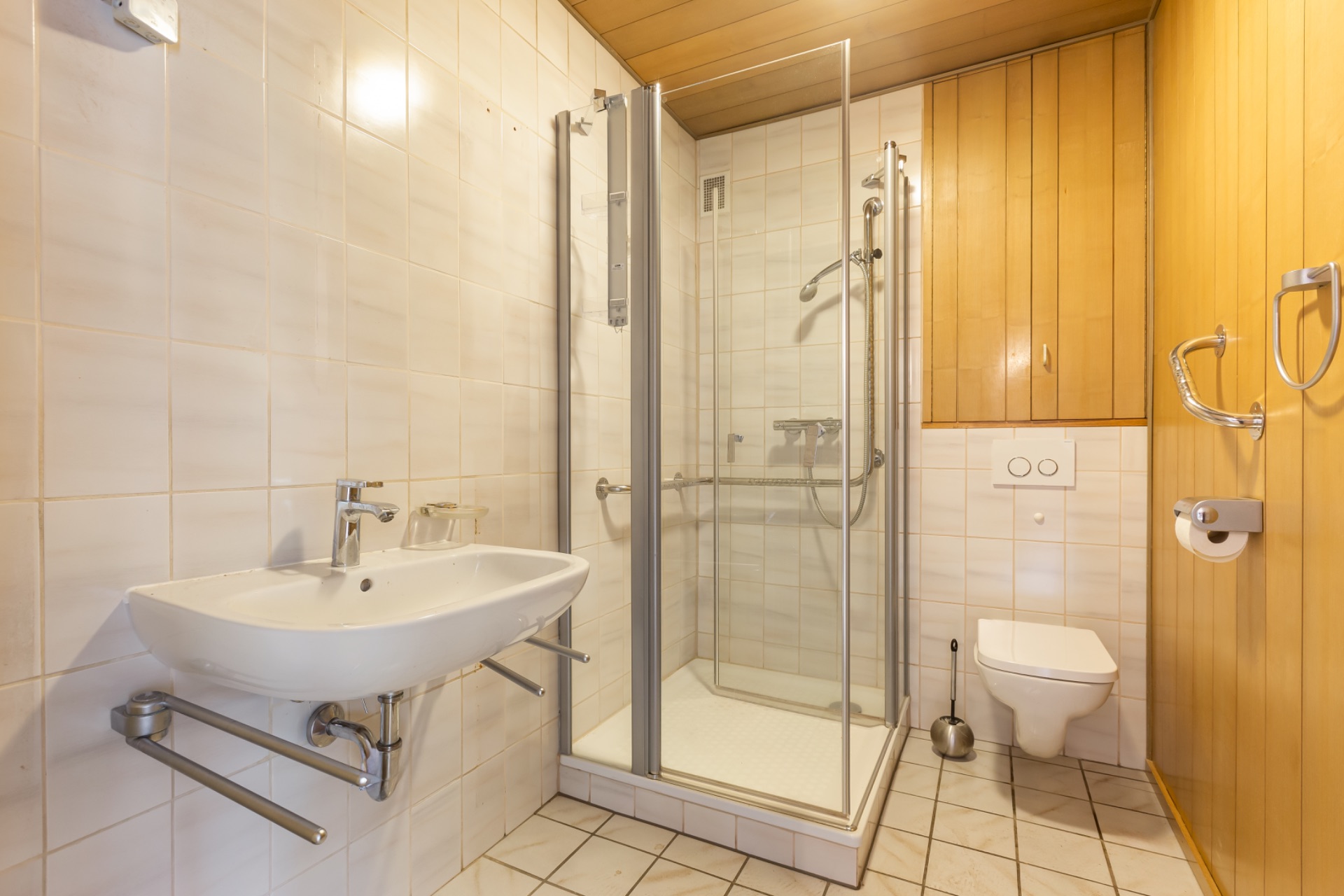 Badezimmer mit Dusche, Waschbecken und Wandtoilette, Holzverkleidung an der rechten Wand.