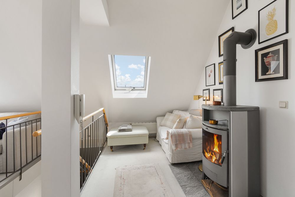 Heller Wohnraum mit Sofa, Kaminofen, Dachfenster und gerahmten Bildern an der Wand.