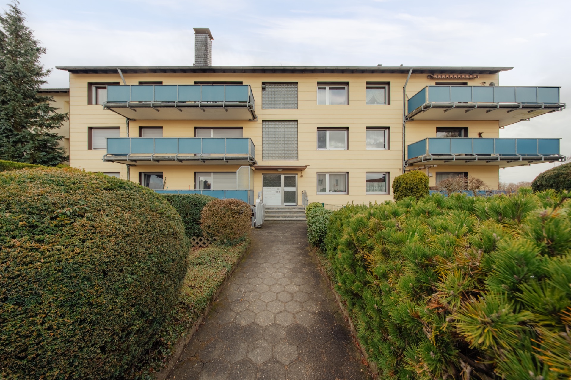 Dreistöckiges Wohnhaus mit mehreren Balkonen, gepflegtem Garten und gepflastertem Weg zum Eingang.