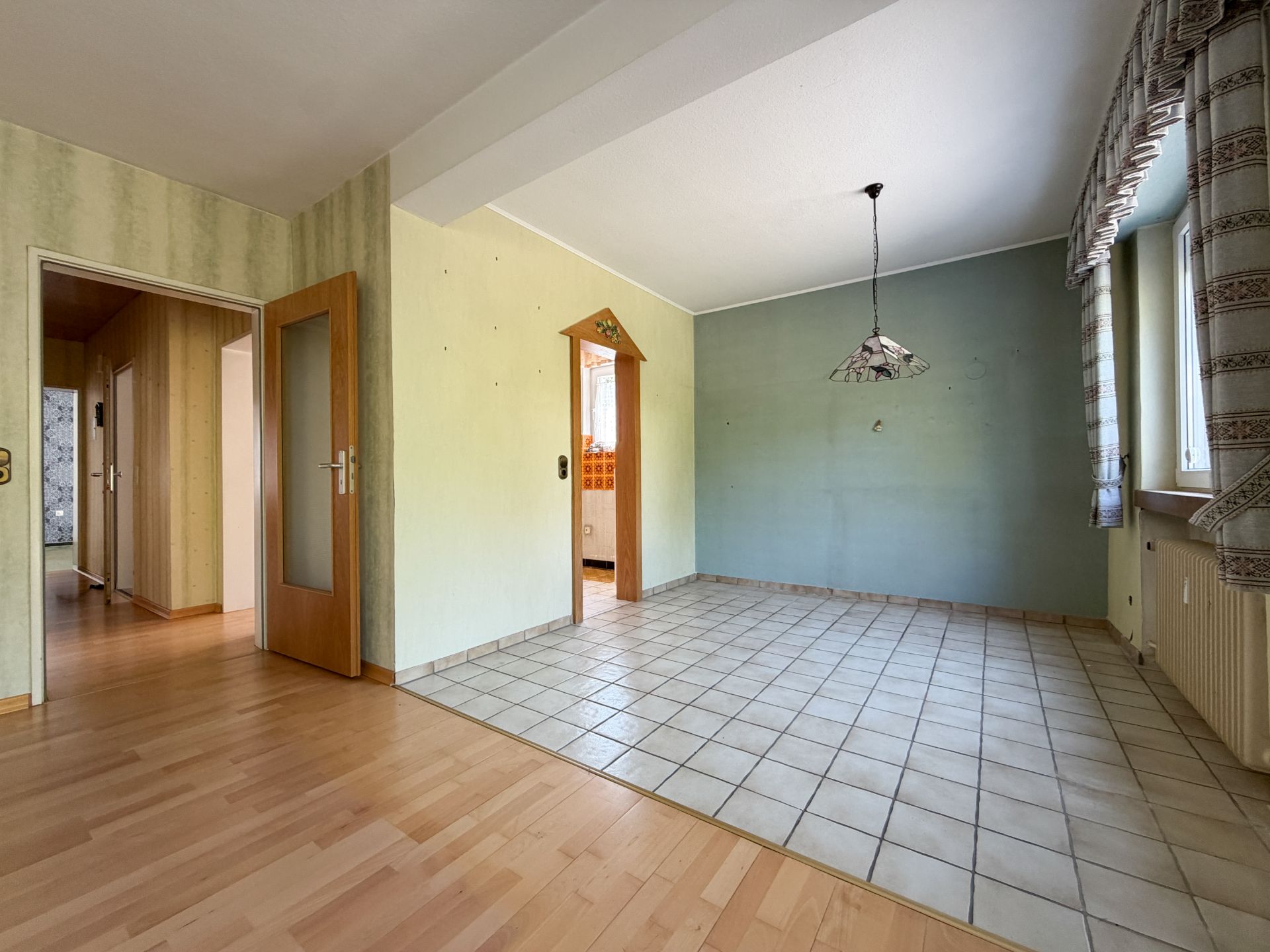 Helles Zimmer mit Fliesen- und Laminatboden, Lampe, Türen und Fenstern mit Vorhängen.