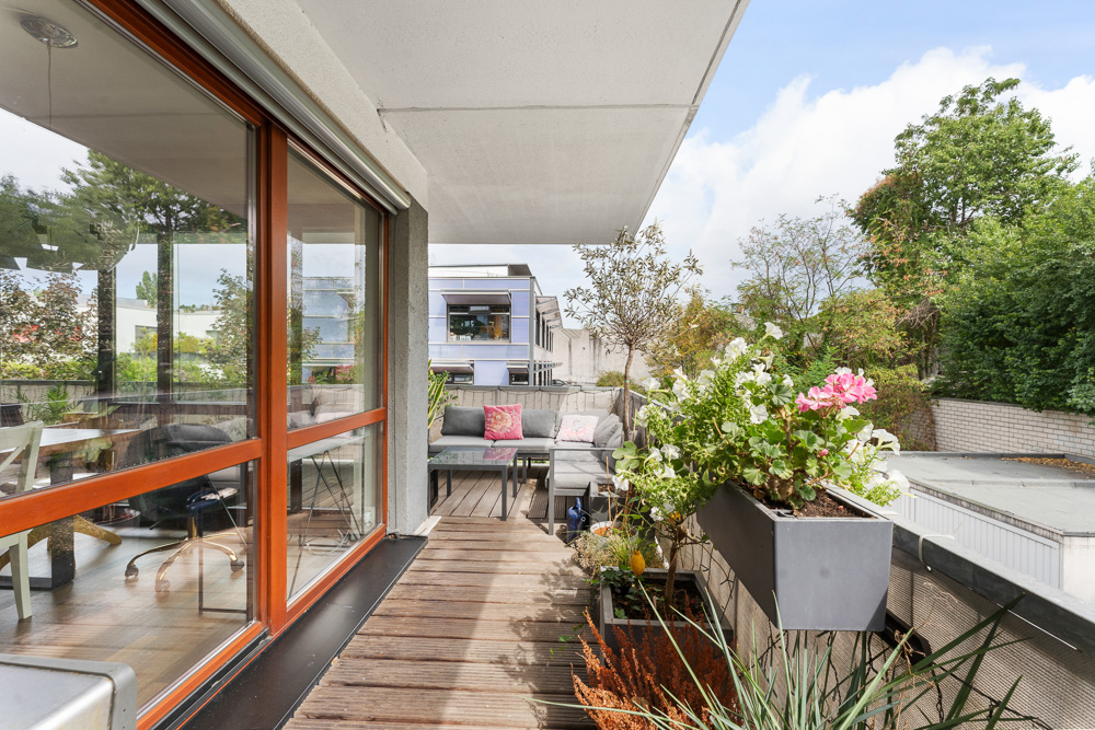 Balkon mit Holzboden, Blumenkästen, Sofa und Tisch, Blick auf Bäume und moderne Wohnhäuser.