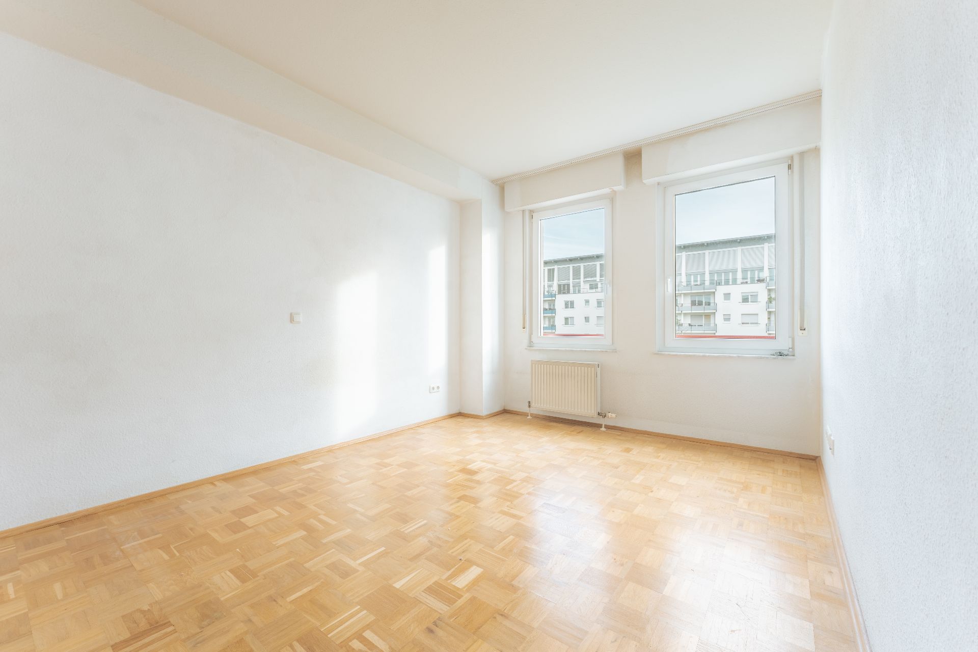 Heller, leerer Raum mit Parkettboden, zwei Fenstern und einem Heizkörper an der Wand.