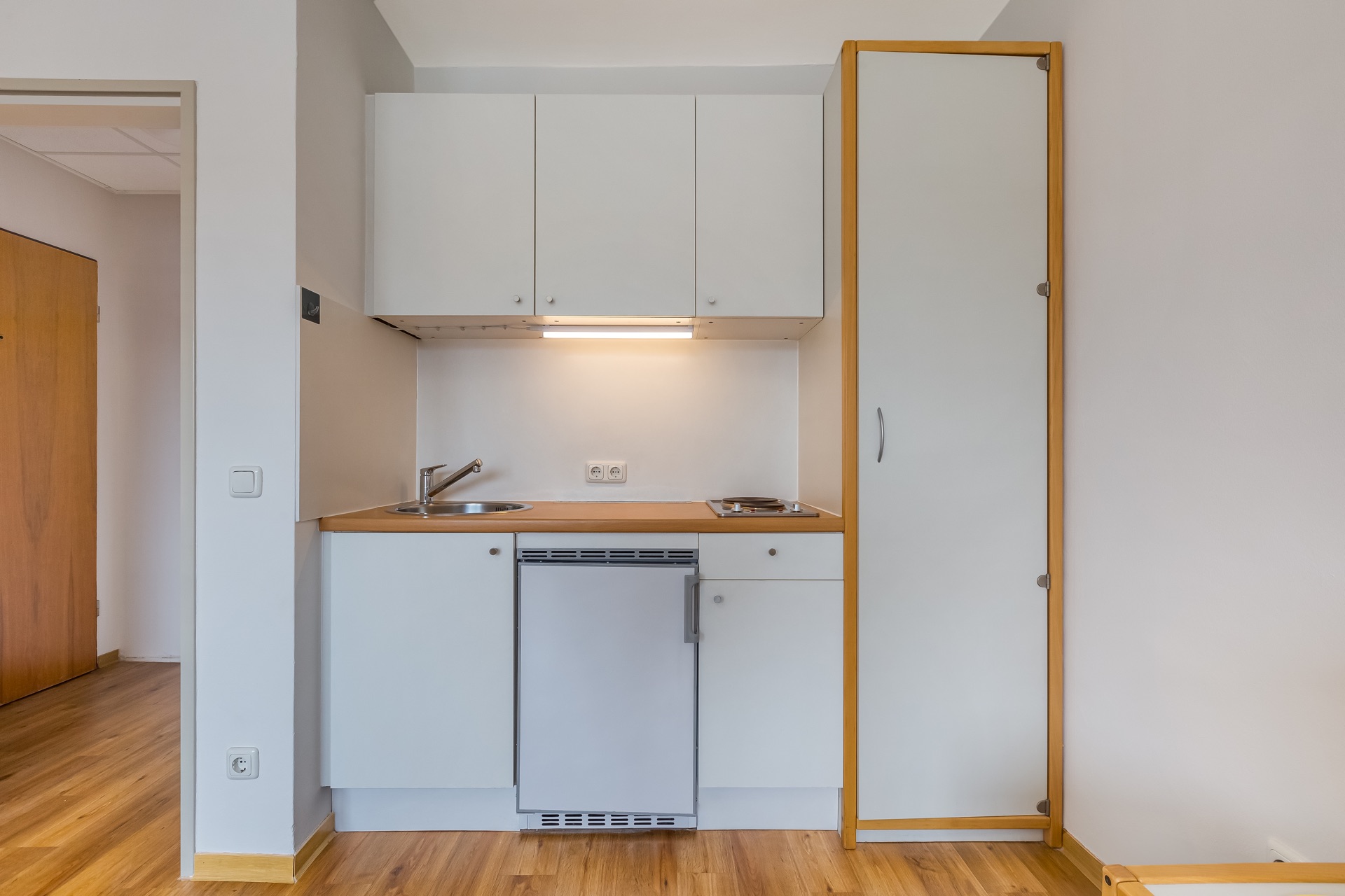 Kleine Küchenzeile mit Spüle, Kochfeld, Kühlschrank und Hängeschränken, Holz- und Weißoptik.