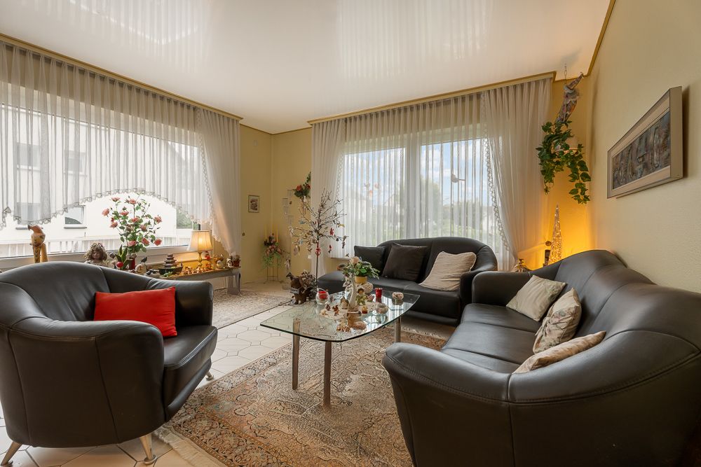 Wohnzimmer mit drei schwarzen Ledersofas, Glastisch, großen Fenstern und Vorhängen.