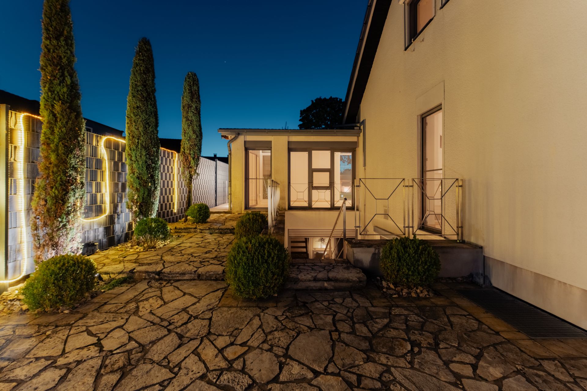Gepflasterter Hof mit kleinen Büschen und Bäumen neben einem Wohnhaus am Abend.