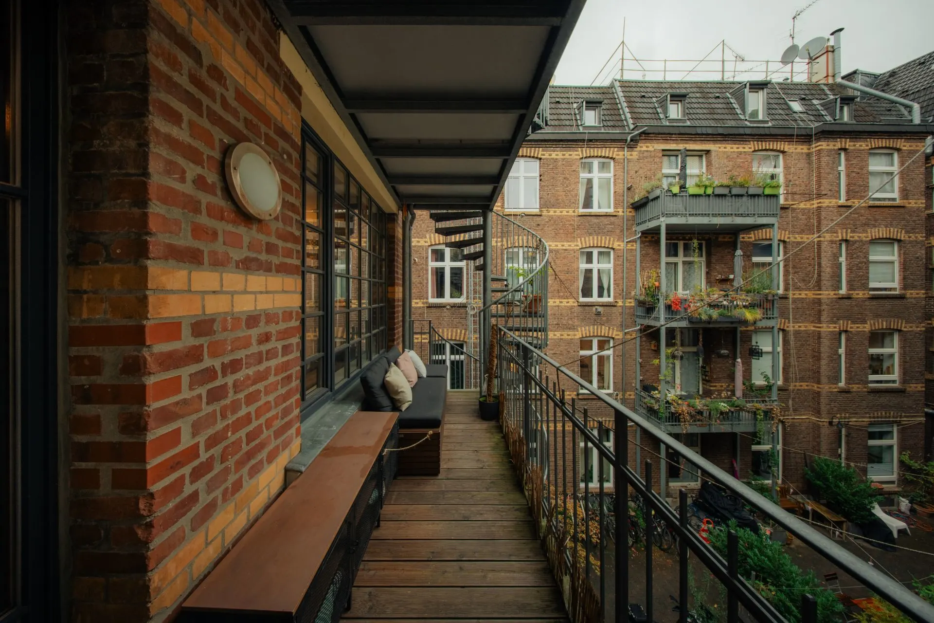 Überdachter Balkon mit Sitzbank und Blick auf einen begrünten Innenhof mit Backsteingebäuden.