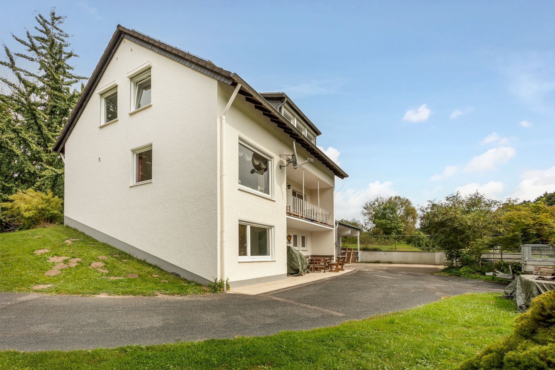 Weißes Wohnhaus mit Balkon, Terrasse und Garten, umgeben von Bäumen und einer Einfahrt.