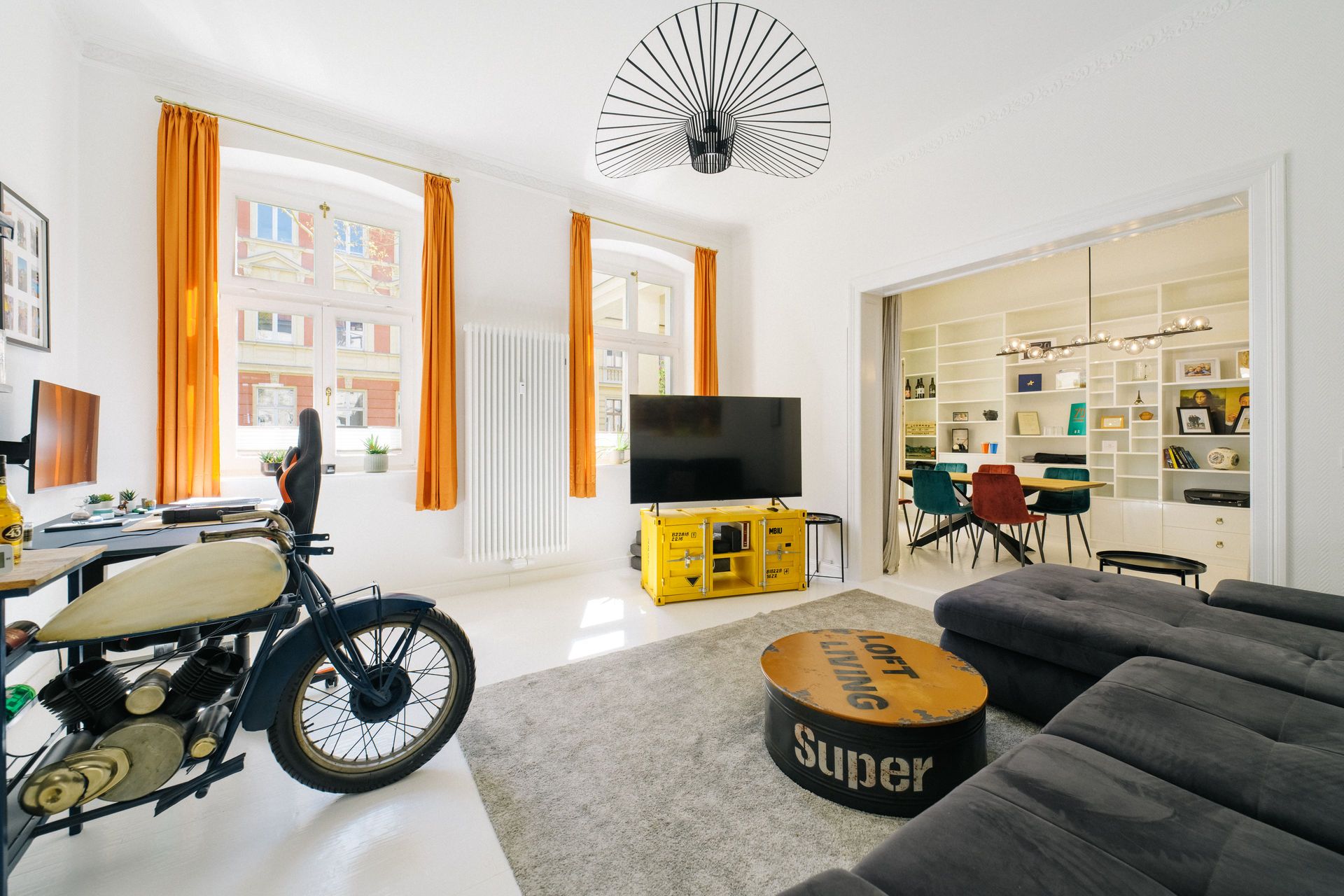 Helles Wohnzimmer mit Sofa, Fernseher, Schreibtisch, Motorrad und orangenen Vorhängen.