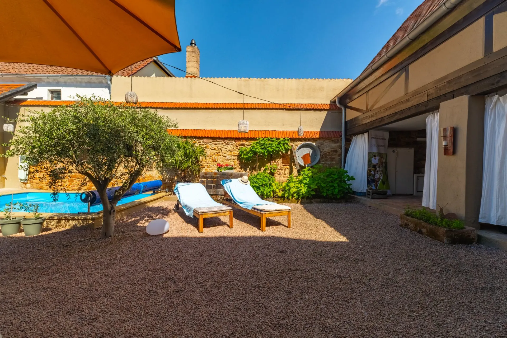 Innenhof mit zwei Liegestühlen, Baum, Sonnenschirm und kleinem Pool auf Kiesboden.