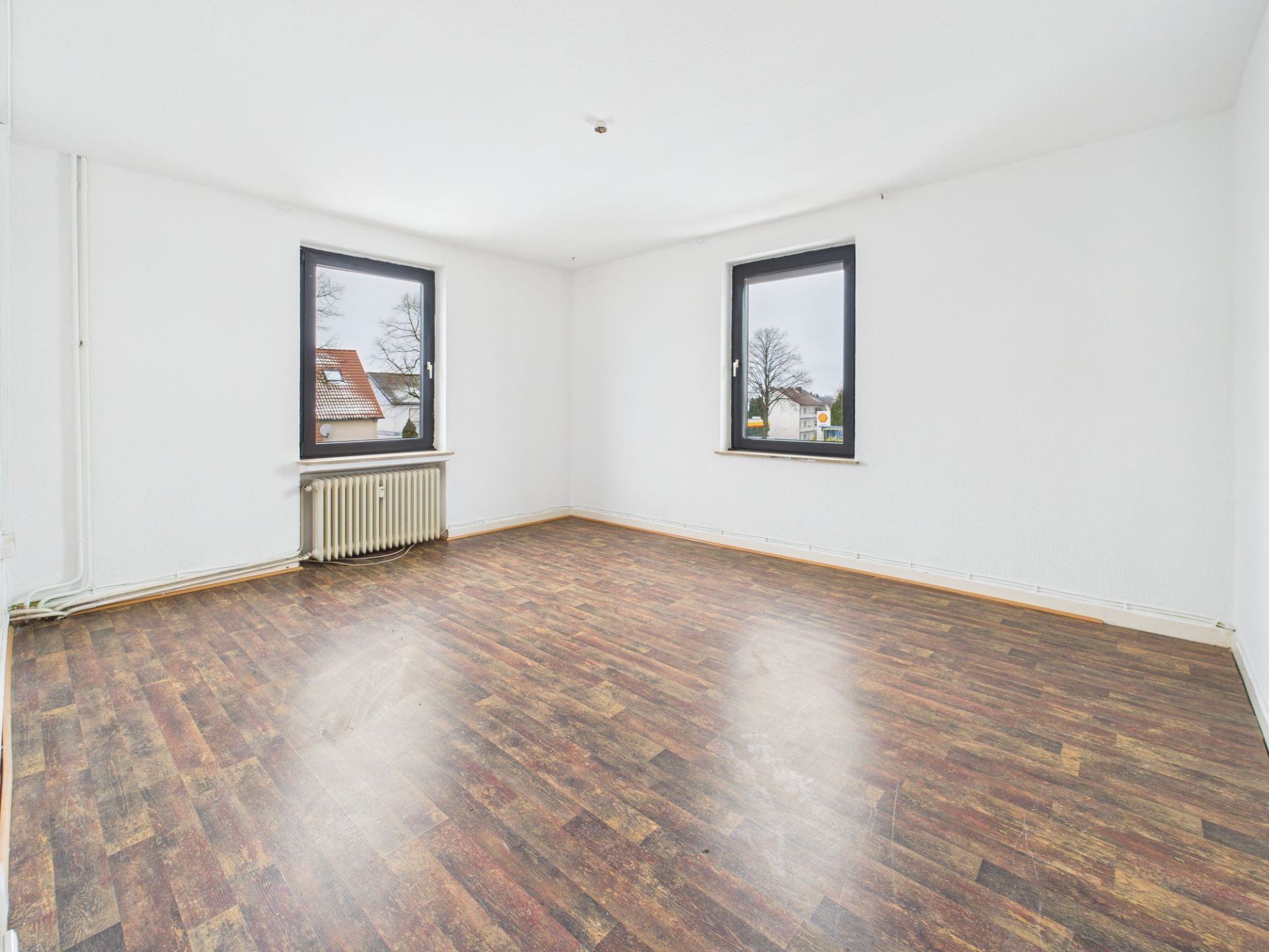 Leerer Raum mit dunklem Holzfußboden, zwei Fenstern und einem Heizkörper unter einem Fenster.