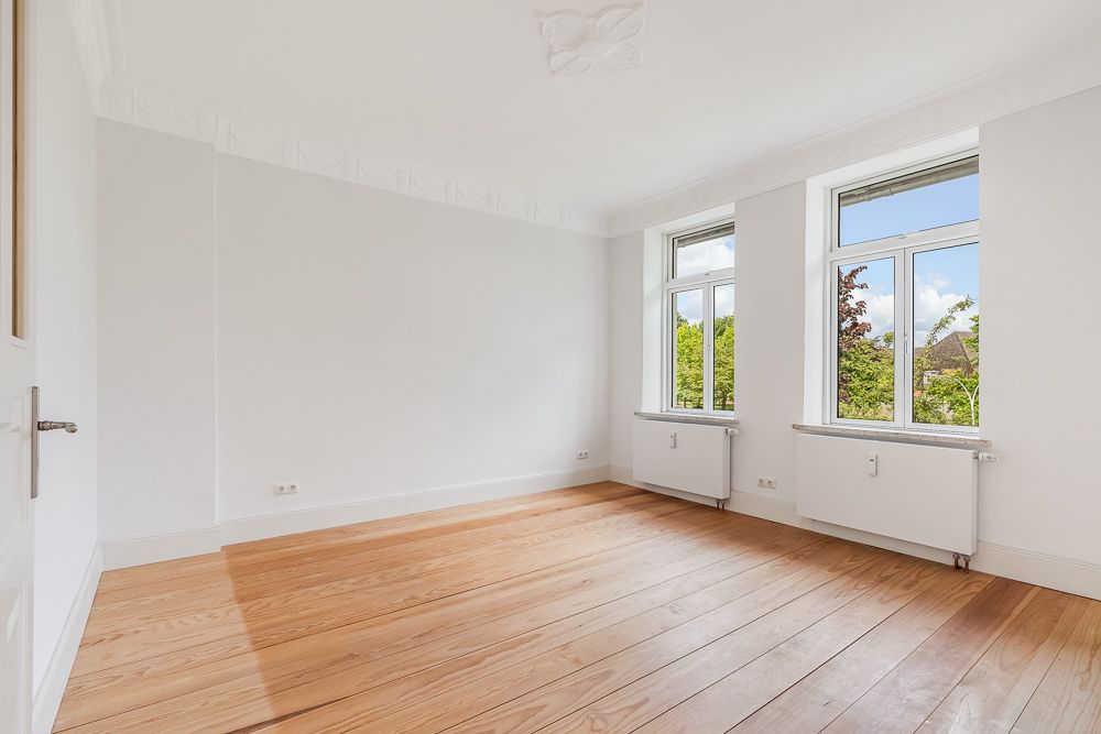 Heller, leerer Raum mit Holzboden und zwei großen Fenstern, durch die Bäume sichtbar sind.